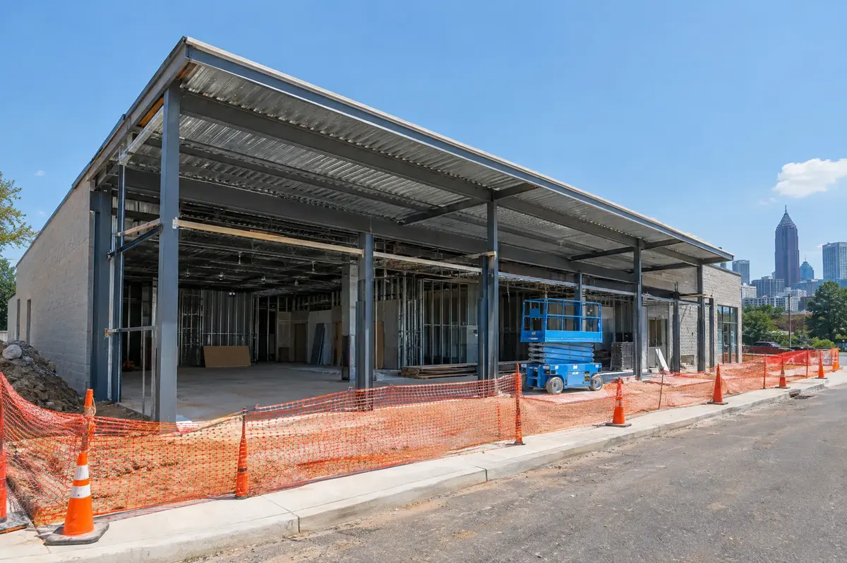Steel framed commercial shell building with a metal roof deck, a scissor lift inside and orange safety fence along the street.