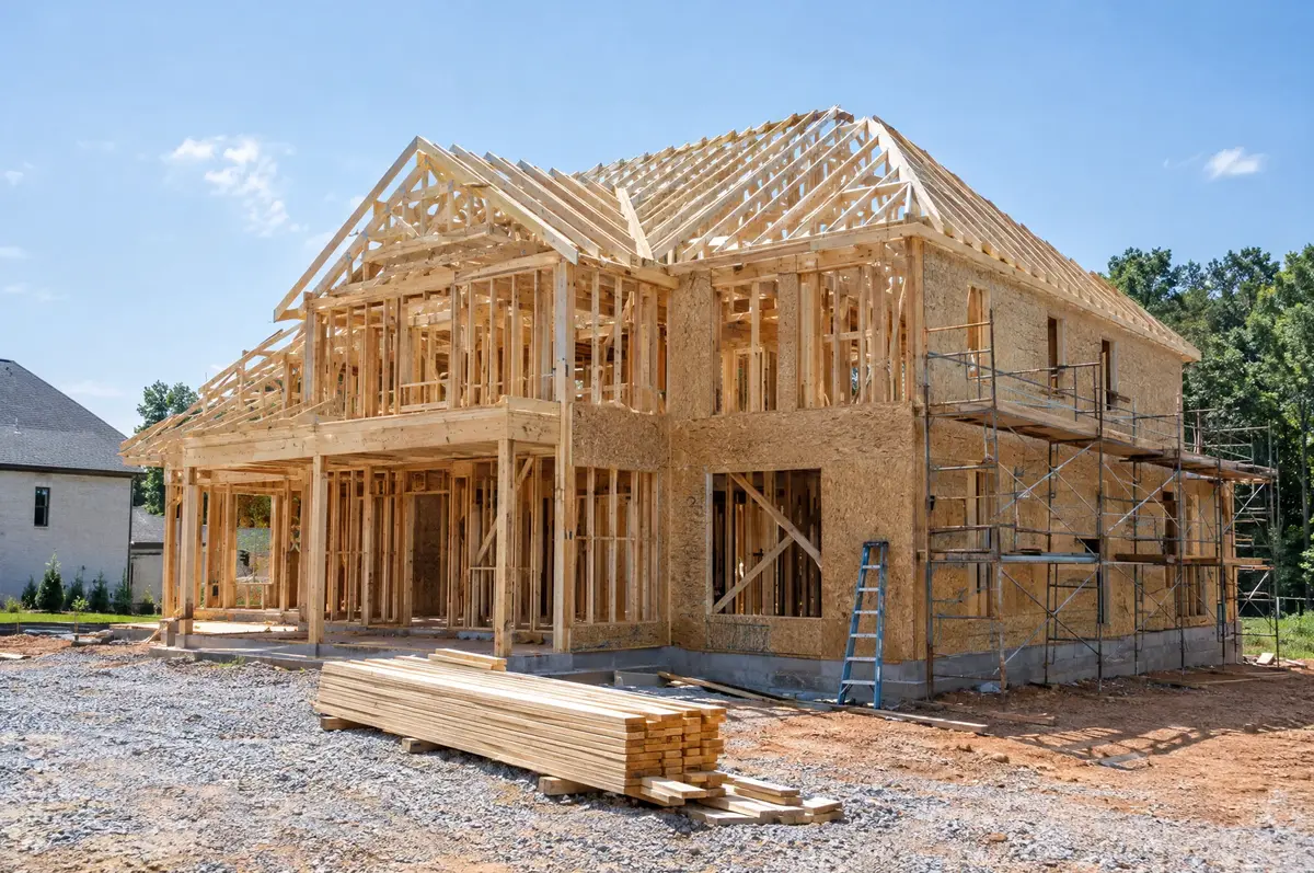 Two storey house under construction with timber framing, roof trusses, scaffolding and a stack of lumber out front.